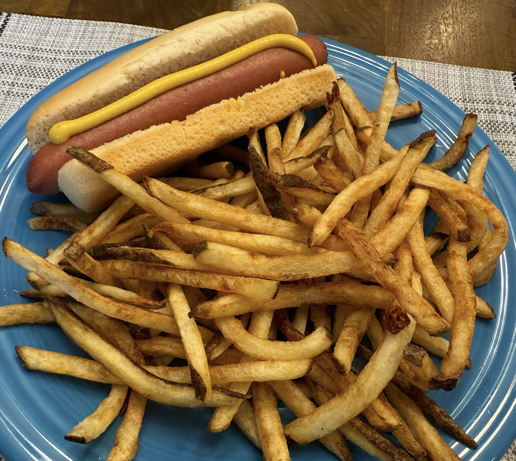 A hot dog with mustard in a bun is served on a blue plate alongside a large pile of golden-brown French fries.