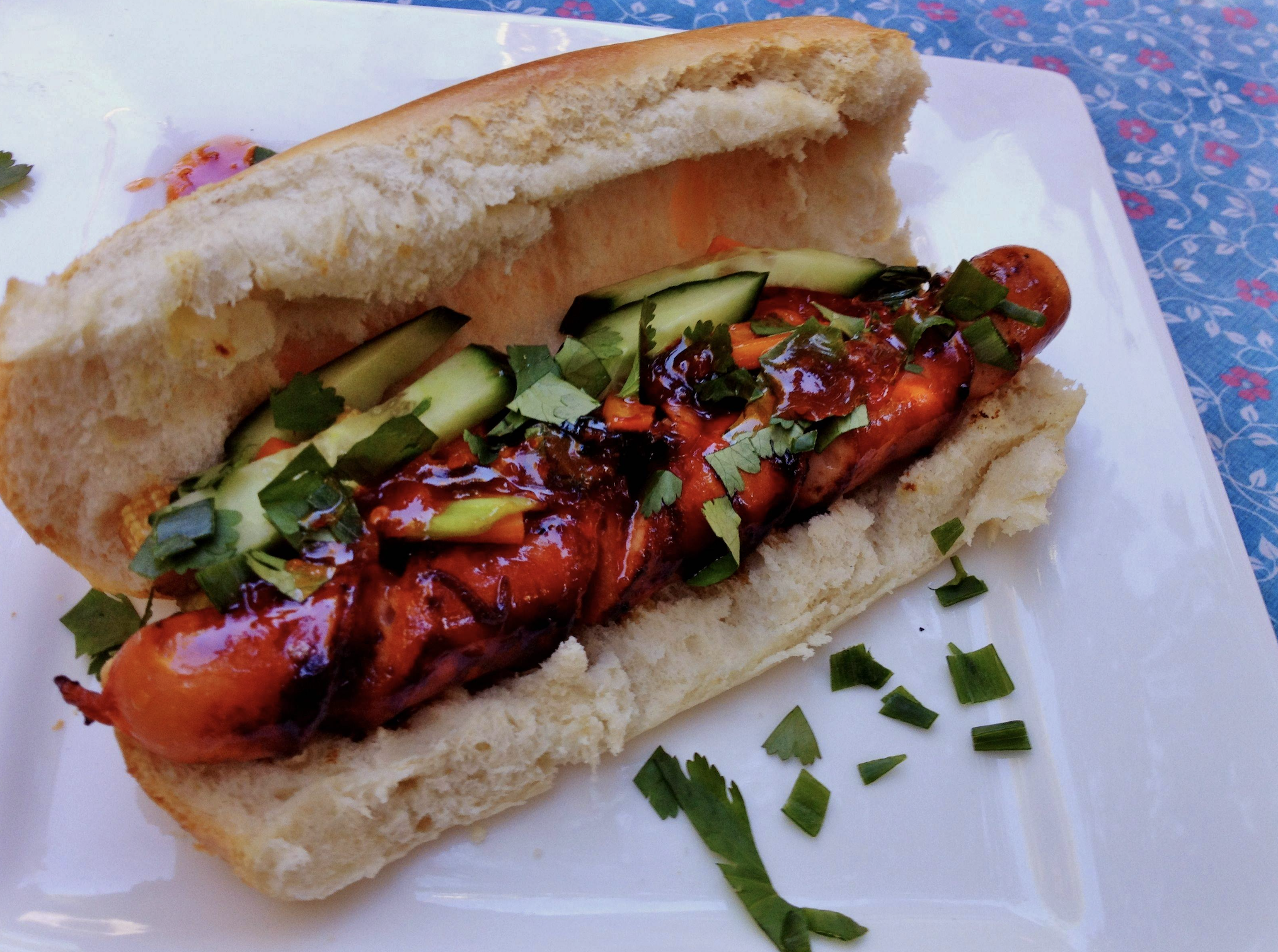 A grilled hot dog in a bun topped with sliced cucumbers, chopped cilantro, and a dark sauce, served on a white plate with a few herb pieces scattered around.