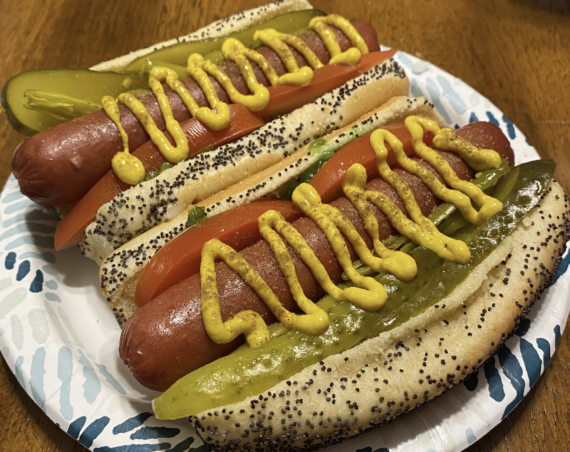 Two Chicago-style hot dogs in poppy seed buns topped with mustard, tomato slices, pickles, and sport peppers, served on a patterned paper plate on a wooden table.