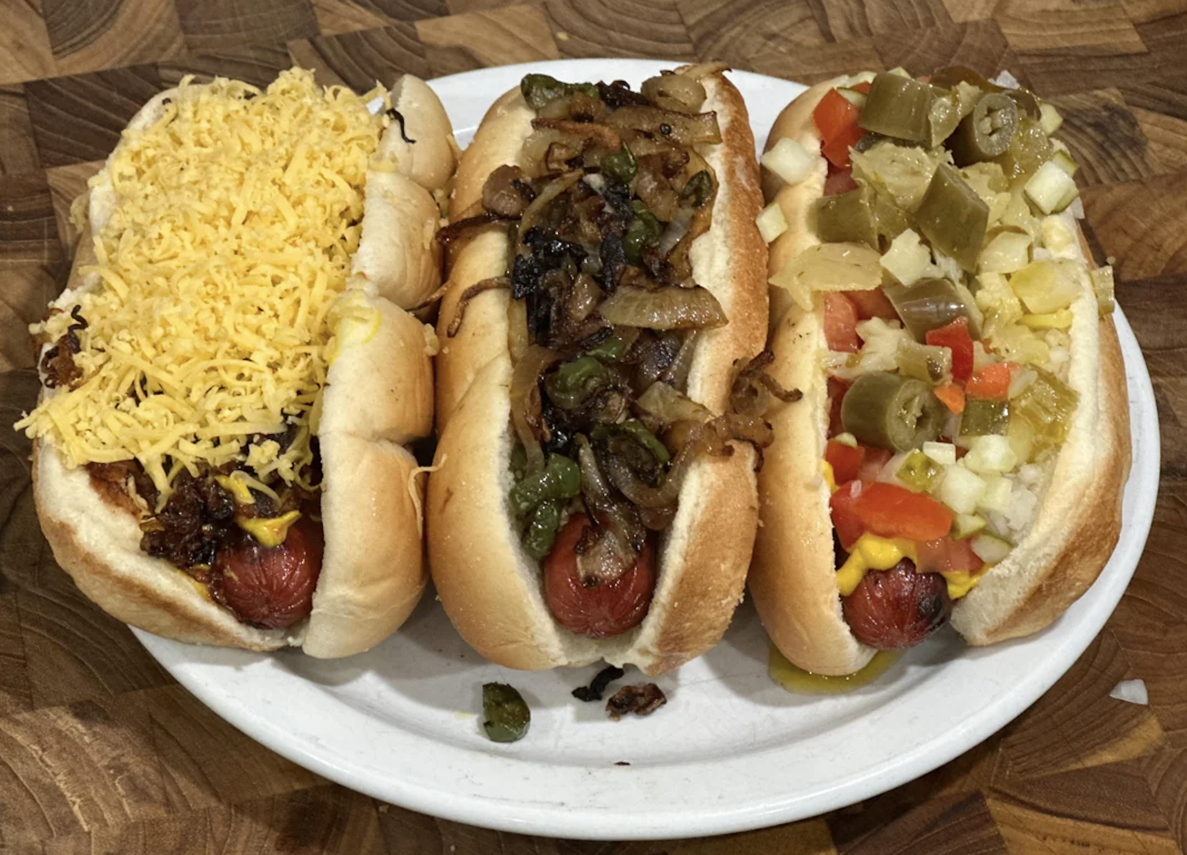 Three hot dogs on a white plate: one with shredded cheese, one with grilled onions and peppers, and one with chopped onions, tomatoes, mustard, and pickled peppers.
