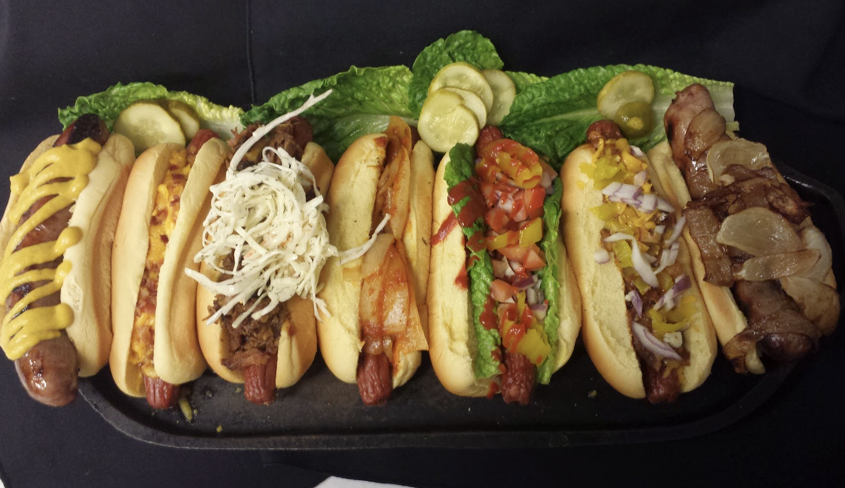 Seven hot dogs in buns, each topped with different ingredients like mustard, cheese, shredded cabbage, pickles, onions, tomatoes, peppers, and grilled onions, are arranged on a platter with lettuce and pickle slices.