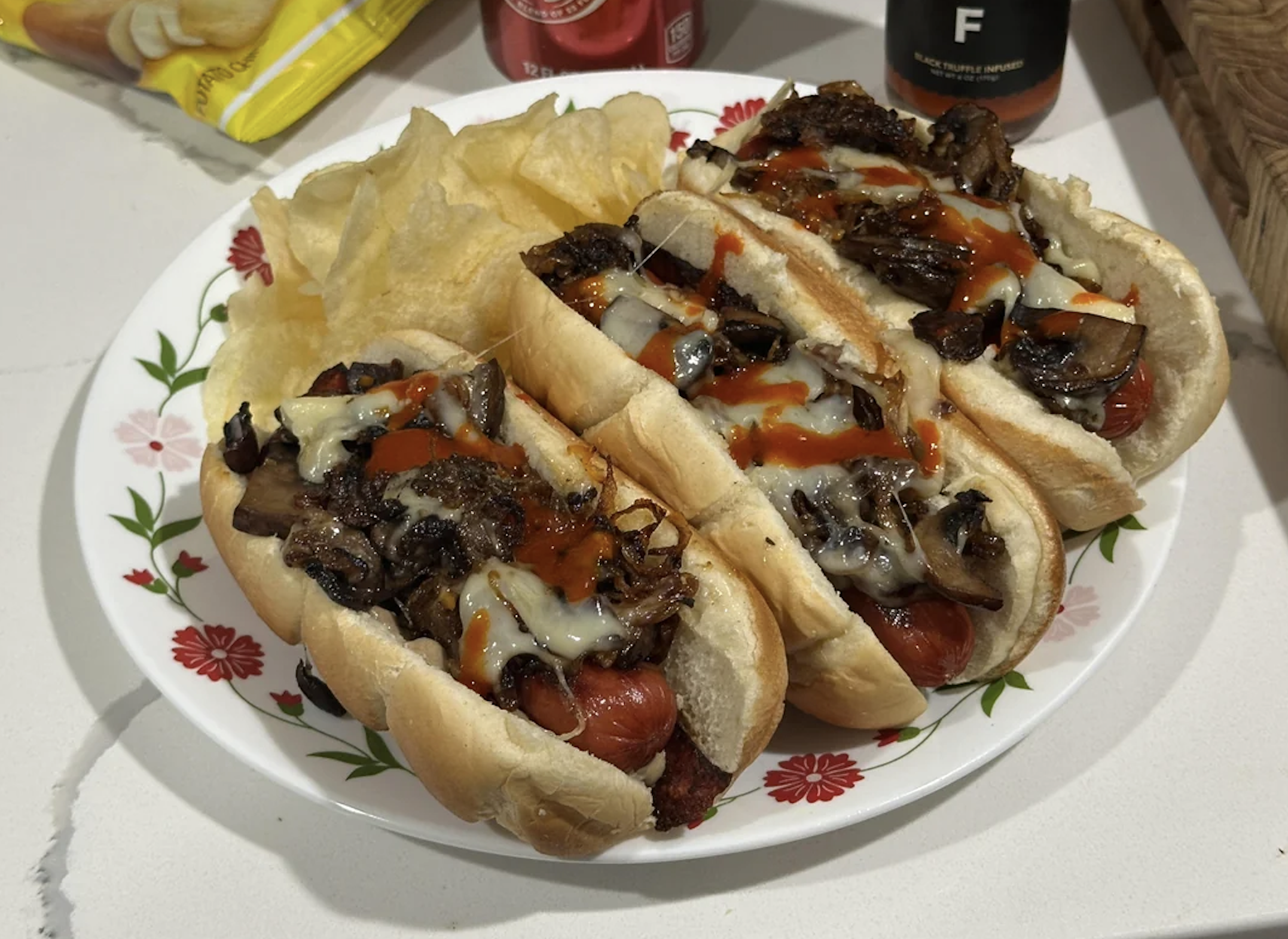 A plate with three loaded hot dogs topped with sautéed mushrooms, melted cheese, and hot sauce, served with a side of potato chips.