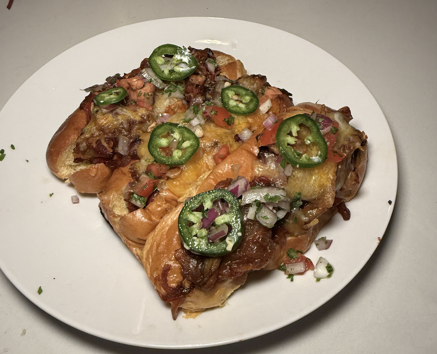 Cheesy pulled pork sliders topped with sliced jalapeños, diced red onions, tomatoes, and herbs on a white plate.