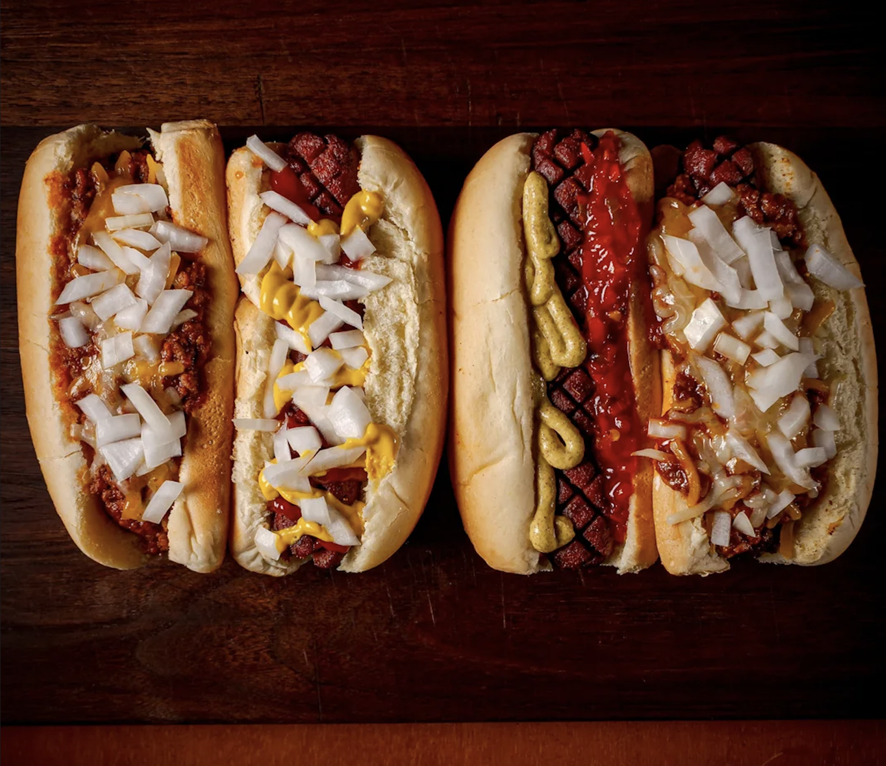 Four hot dogs on a wooden surface, each topped with diced onions and different condiments: chili, mustard, ketchup, and what seems to be a mix of sauces.