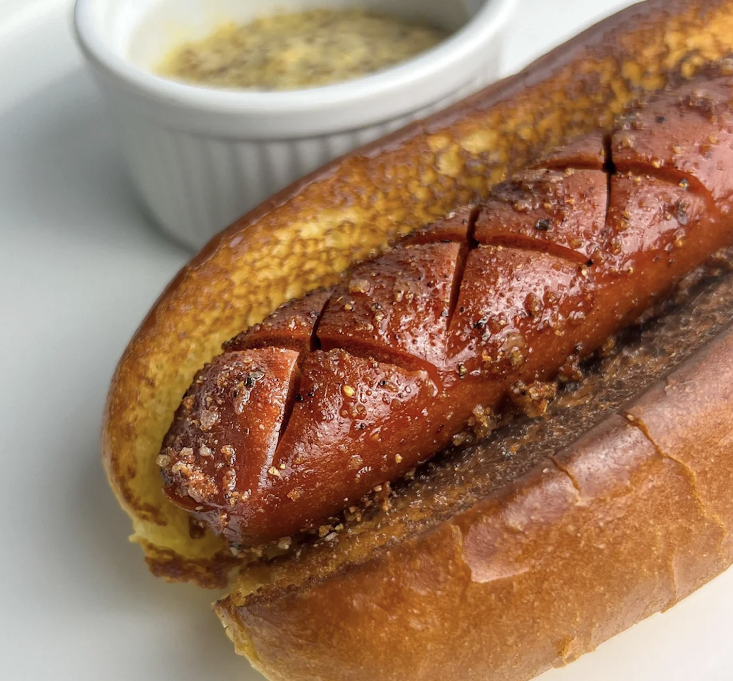 A toasted hot dog bun holds a grilled sausage with crosshatched cuts. A small white ramekin filled with grainy mustard sits in the background on a white plate.