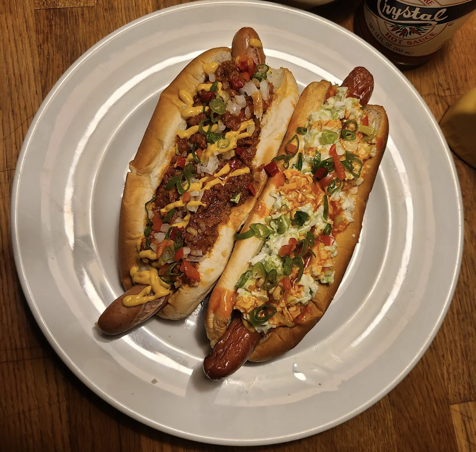 Two hot dogs on a white plate: one topped with chili, onions, and mustard, the other with coleslaw, green onions, jalapeños, and hot sauce. The plate sits on a wooden table.