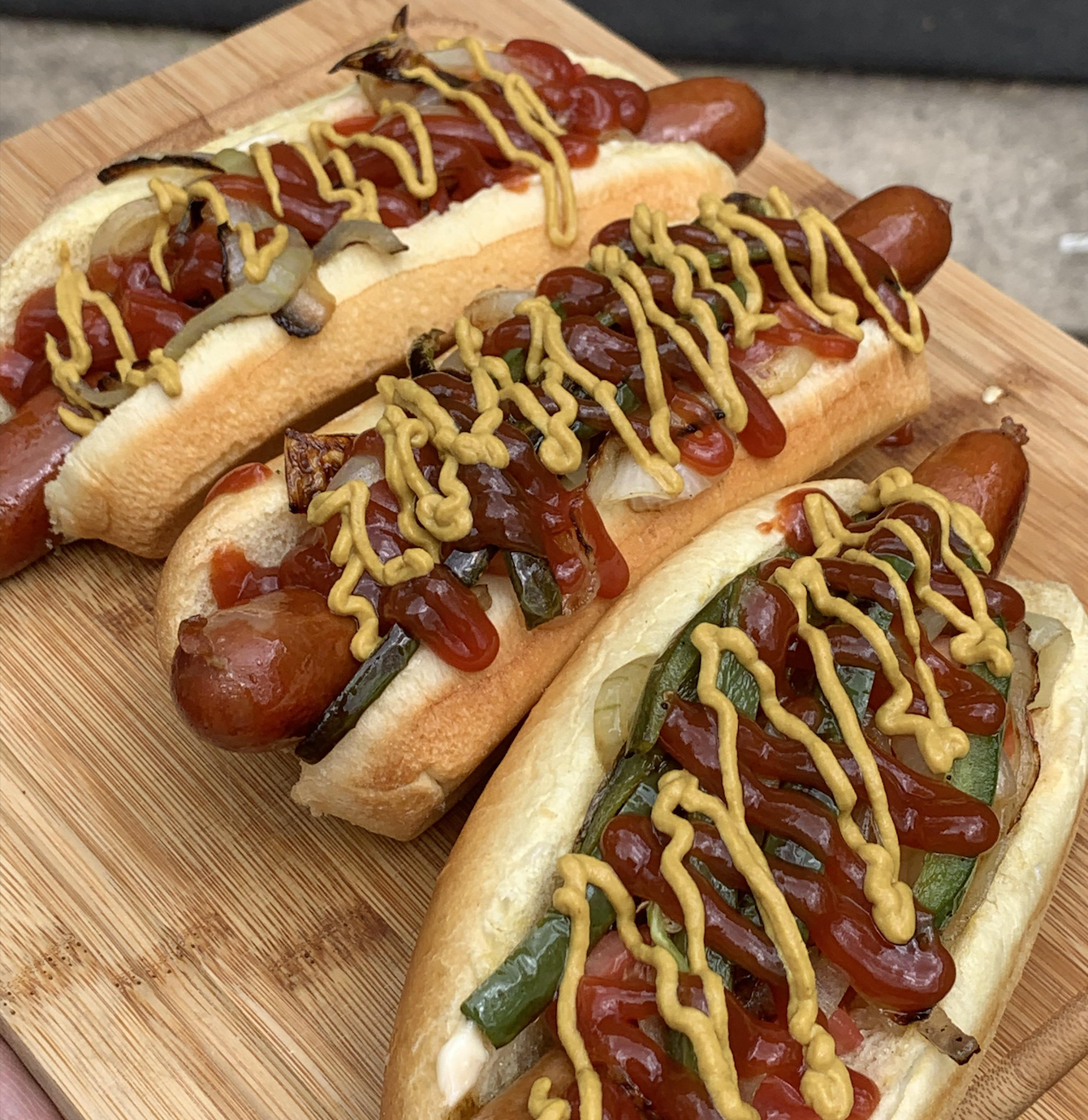 Three grilled hot dogs in buns topped with ketchup, mustard, grilled onions, and green peppers, served on a wooden board.