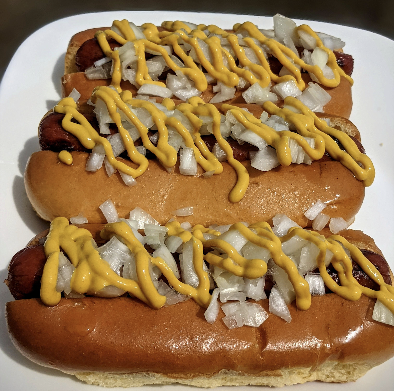 Three grilled hot dogs in buns are topped with chopped onions and zigzag lines of yellow mustard, arranged on a white rectangular plate.