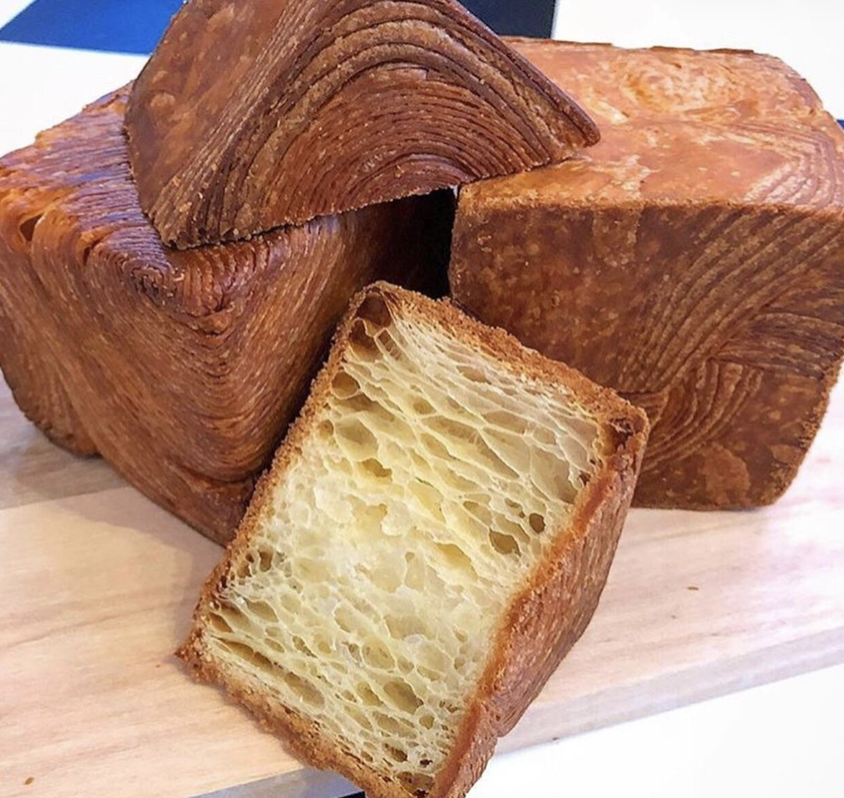 Loaves of cube-shaped pastries with flaky, layered golden-brown crusts and a light, airy interior, displayed on a wooden surface.