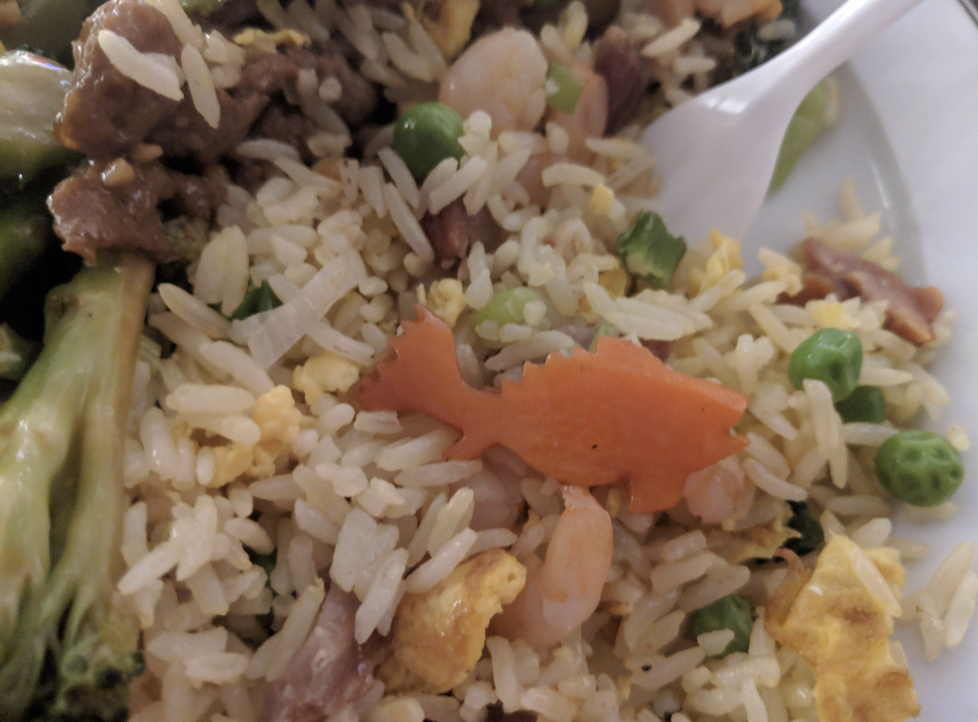 A plate of fried rice with peas, egg, shrimp, and a carrot slice cut into the shape of a fish. A plastic fork and some pieces of broccoli are visible on the side.