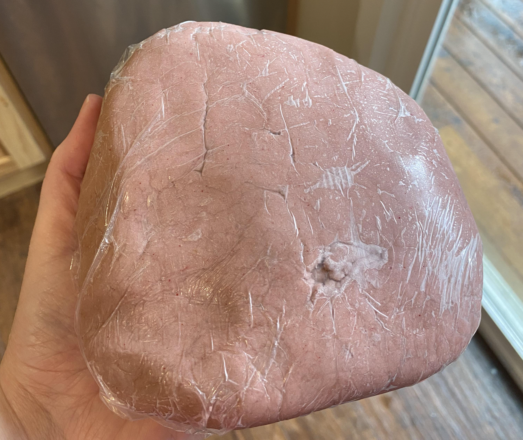 A hand holds a large, lump-shaped piece of pink dough wrapped in clear plastic wrap, with wrinkles and a small indent on its surface.
