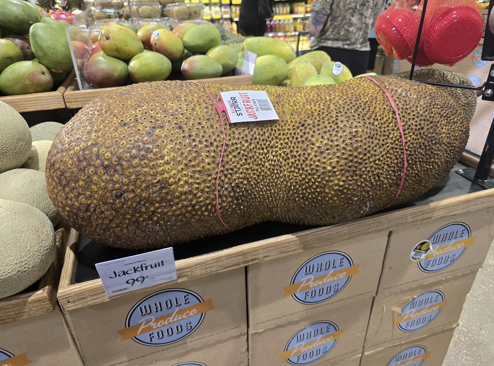 A large, brown, bumpy jackfruit is displayed on top of Whole Foods boxes with a price sign. Mangos and other produce are visible in the background.