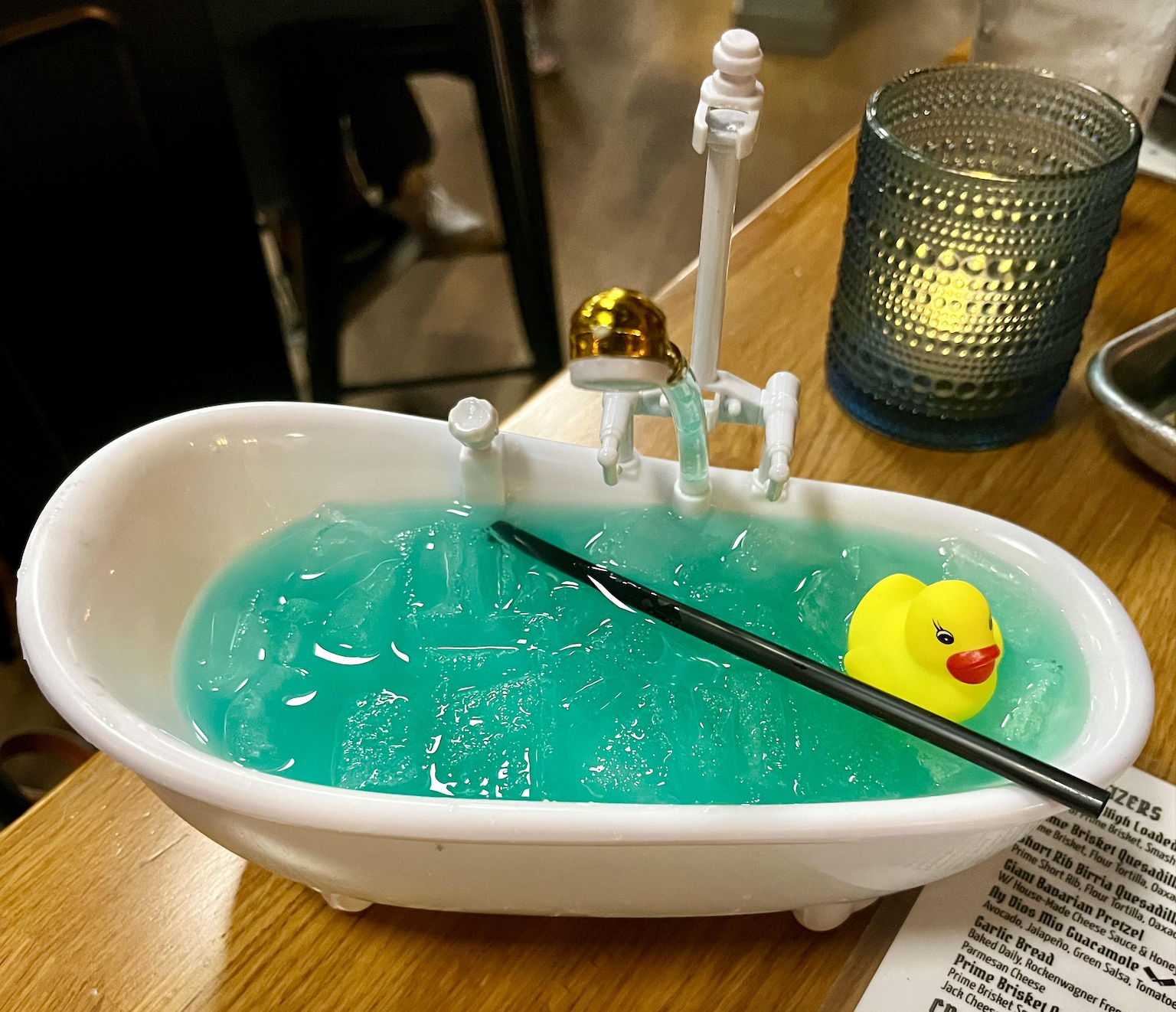 A blue cocktail with ice is served in a miniature bathtub-shaped glass, garnished with a small yellow rubber duck and a black straw. A textured blue glass and a menu are on the wooden table beside the drink.