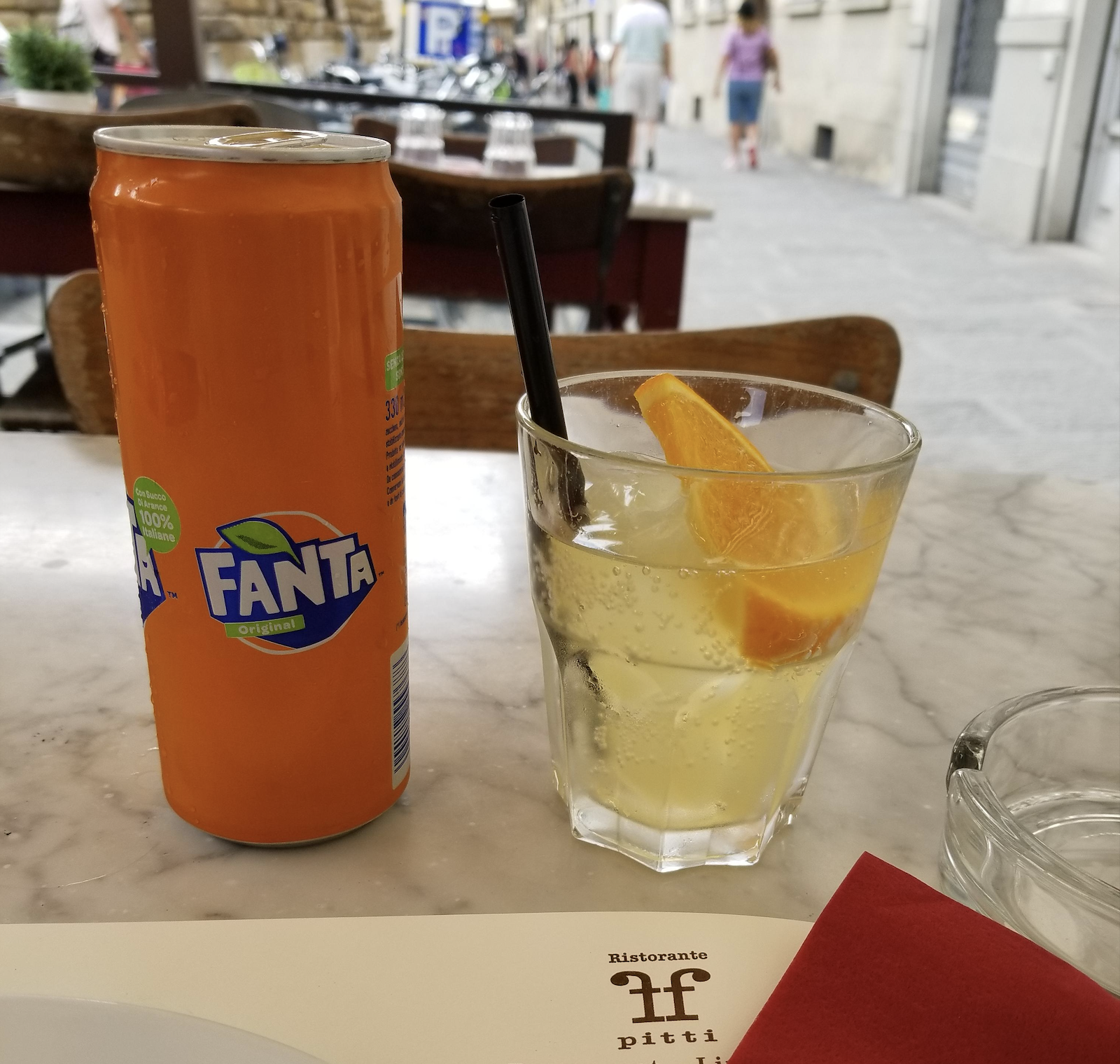 A can of Fanta sits on a marble table next to a glass with ice, orange slices, and a black straw. The setting appears to be an outdoor café, with blurred people and buildings in the background.