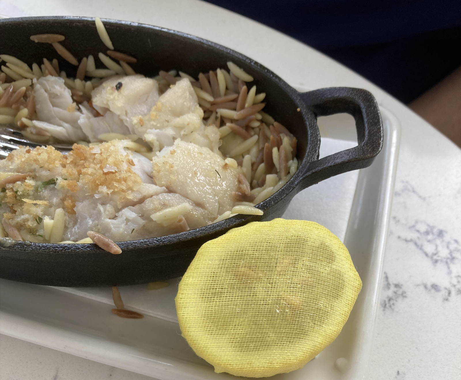 A cast iron dish with white fish and orzo pasta, partially eaten, sits on a plate. A lemon wrapped in yellow mesh is next to the dish.