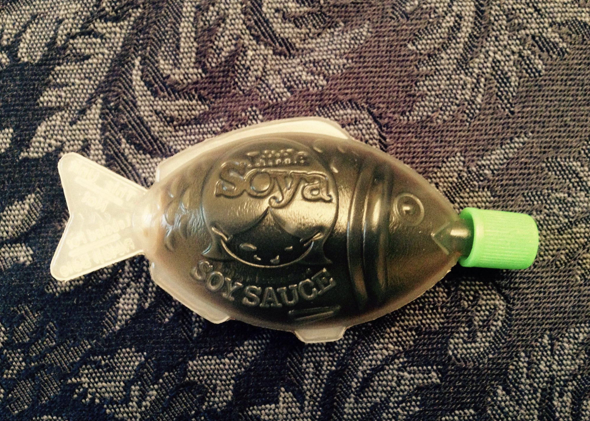 A small, fish-shaped plastic container filled with soy sauce, featuring a green twist cap, rests on a dark, patterned fabric background.