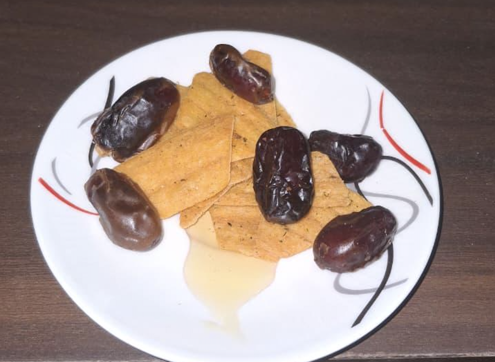 A white plate with rectangular, spiced crackers and several dates, placed on a dark wooden surface. Some syrup is visible beneath the crackers.