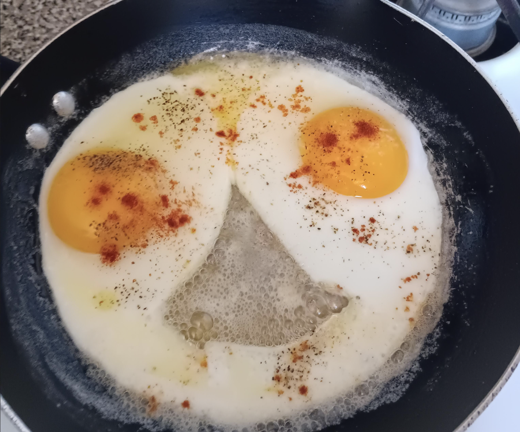 Two sunny-side-up eggs frying in a pan, with the yolks and egg whites resembling a smiling face. Red seasoning is sprinkled on the eggs, and bubbles form in the hot pan.