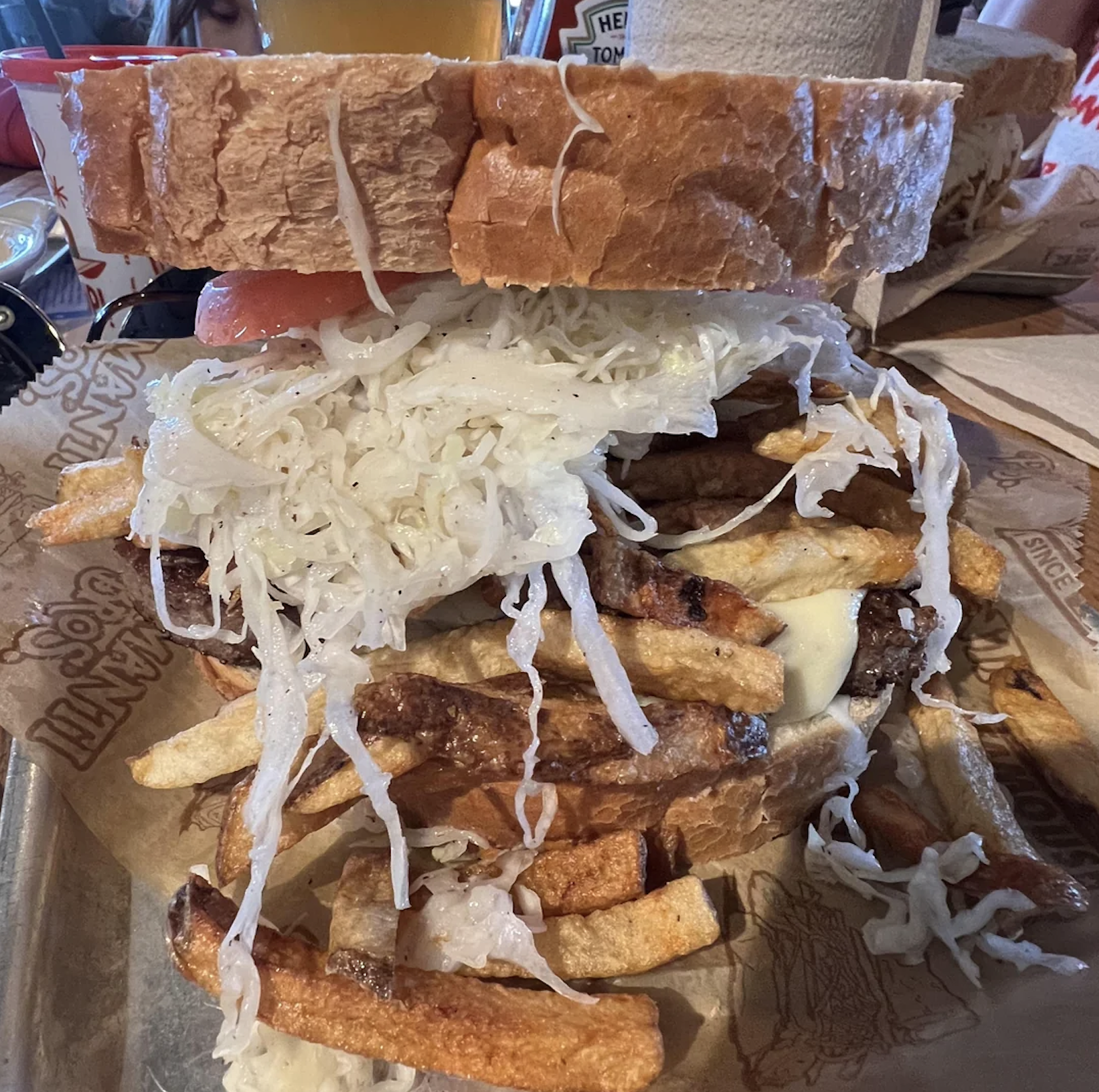 A large sandwich with thick bread slices, piled high with coleslaw, French fries, meat, and cheese, served on a tray with branded paper beneath it.