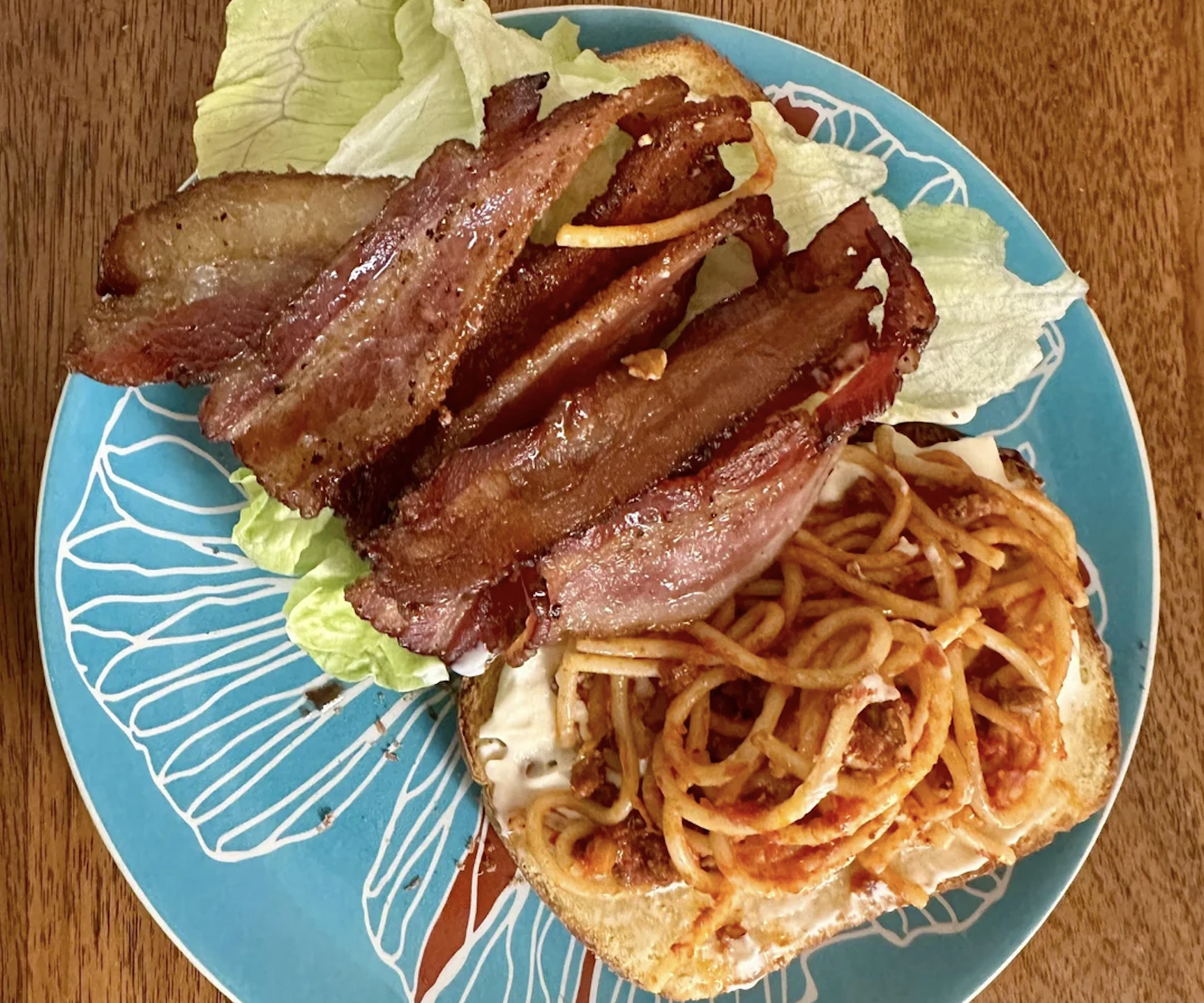 An open-faced sandwich on a blue plate topped with spaghetti, melted cheese, and strips of crispy bacon, with leafy lettuce on the side.