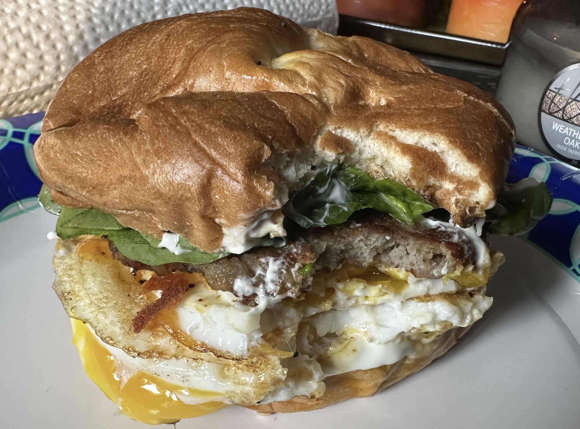A breakfast sandwich with egg, bacon, lettuce, and mayonnaise on a bun, with a large bite taken out of it, sits on a plate.