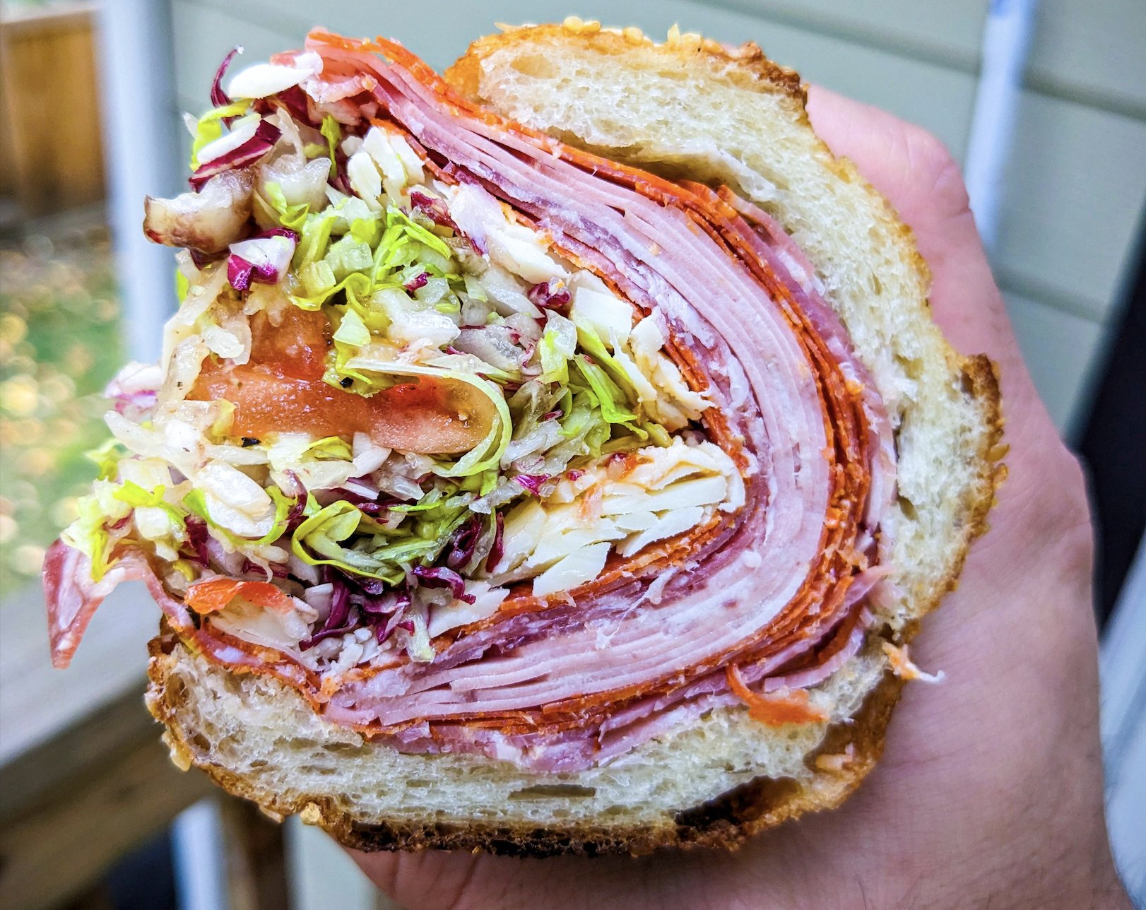 A hand holds a thick sandwich filled with layers of deli meats, shredded lettuce, tomato, and cheese on a sliced roll, shown close up.