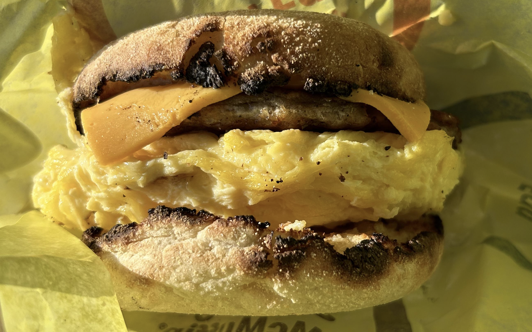 A close-up of a breakfast sandwich with scrambled eggs, cheddar cheese, and sausage on a toasted English muffin, wrapped in yellow paper.