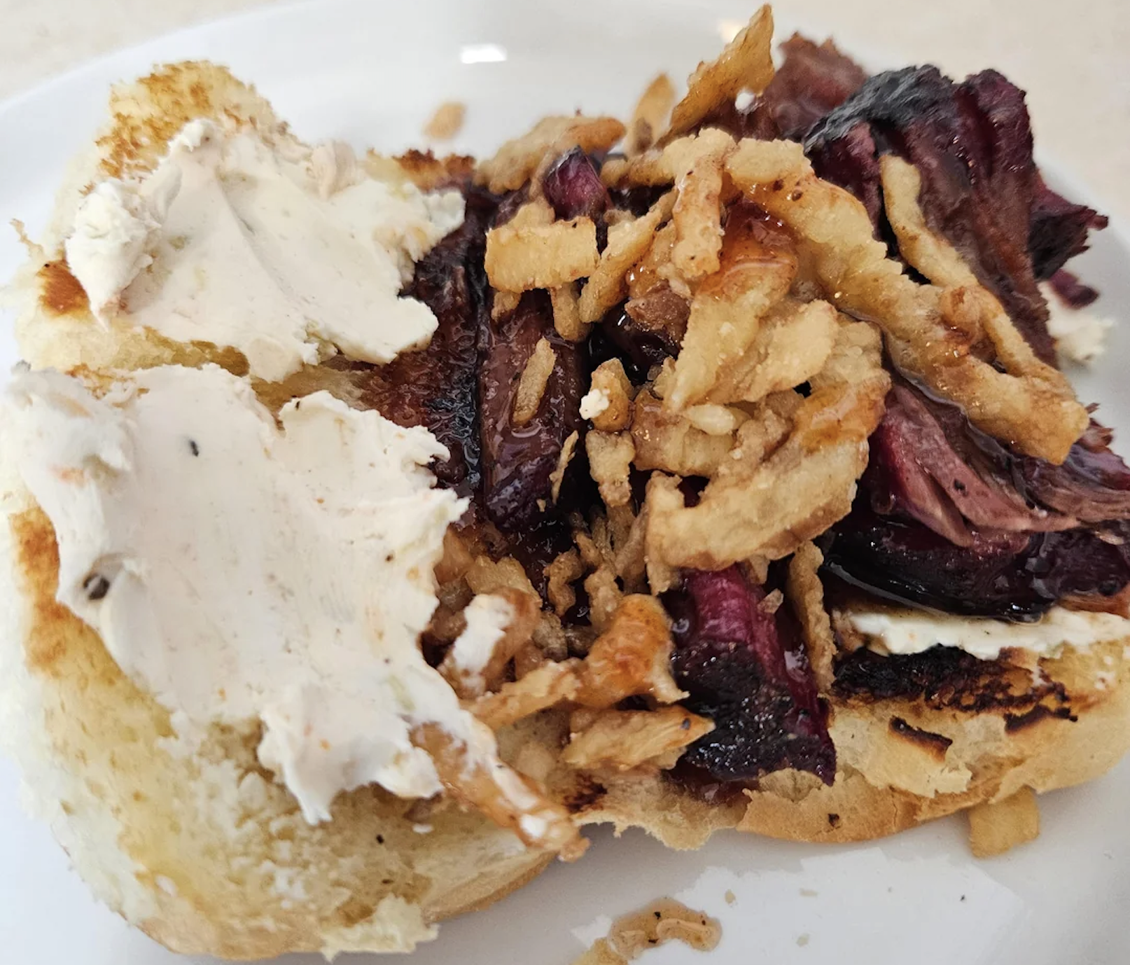 A sandwich with cream cheese, crispy fried onions, and slices of roasted beef on a toasted bun, served on a white plate.