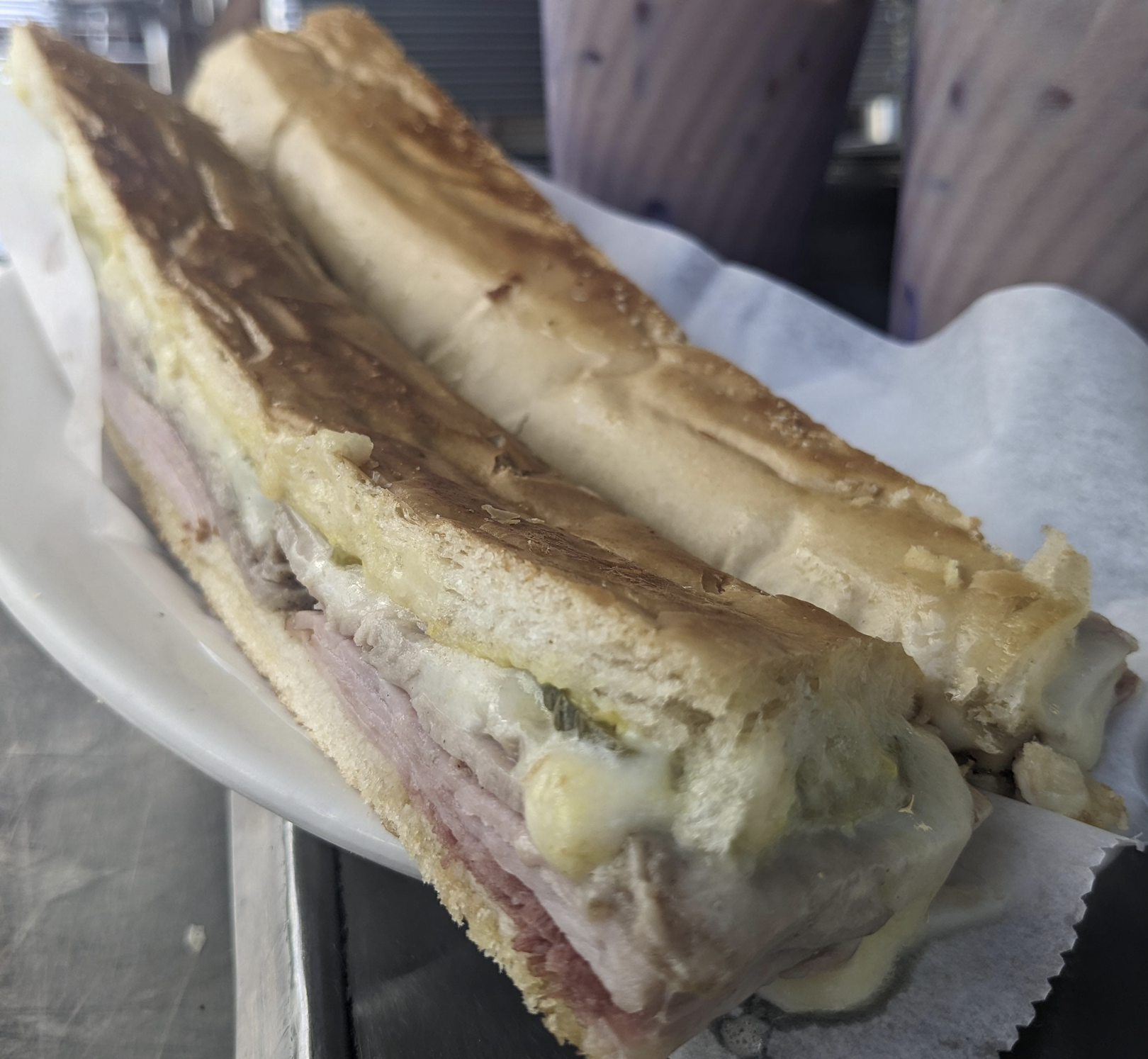 A close-up of a pressed Cuban sandwich with ham, pork, cheese, and pickles on a white plate, with two cups of a purple drink in the background.