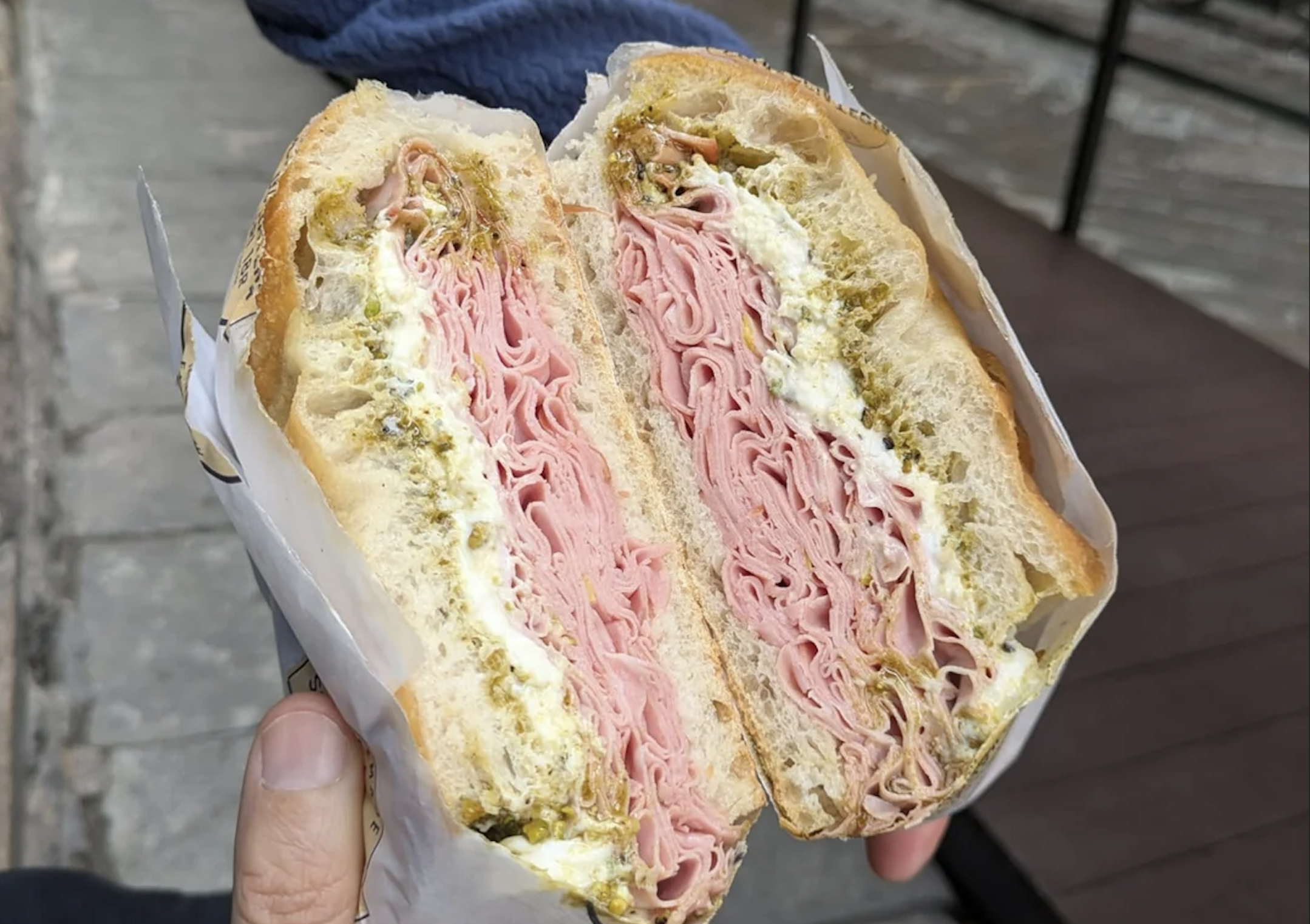A hand holds a large sandwich cut in half, filled with thinly sliced deli meat, cheese, and green relish or sauce, on crusty bread with a soft interior. The background features a stone sidewalk and wooden deck.