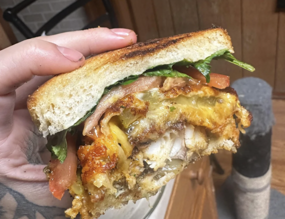 A hand holds a thick sandwich filled with fried chicken, melted cheese, lettuce, and tomato between toasted bread slices.