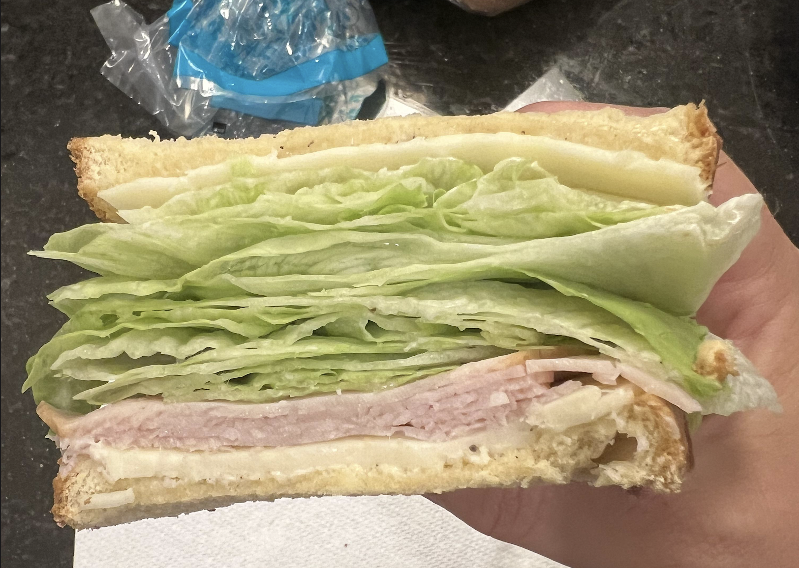 A hand holds a sandwich with layers of turkey, Swiss cheese, and several large leaves of lettuce between slices of white bread. A plastic bag and countertop are visible in the background.