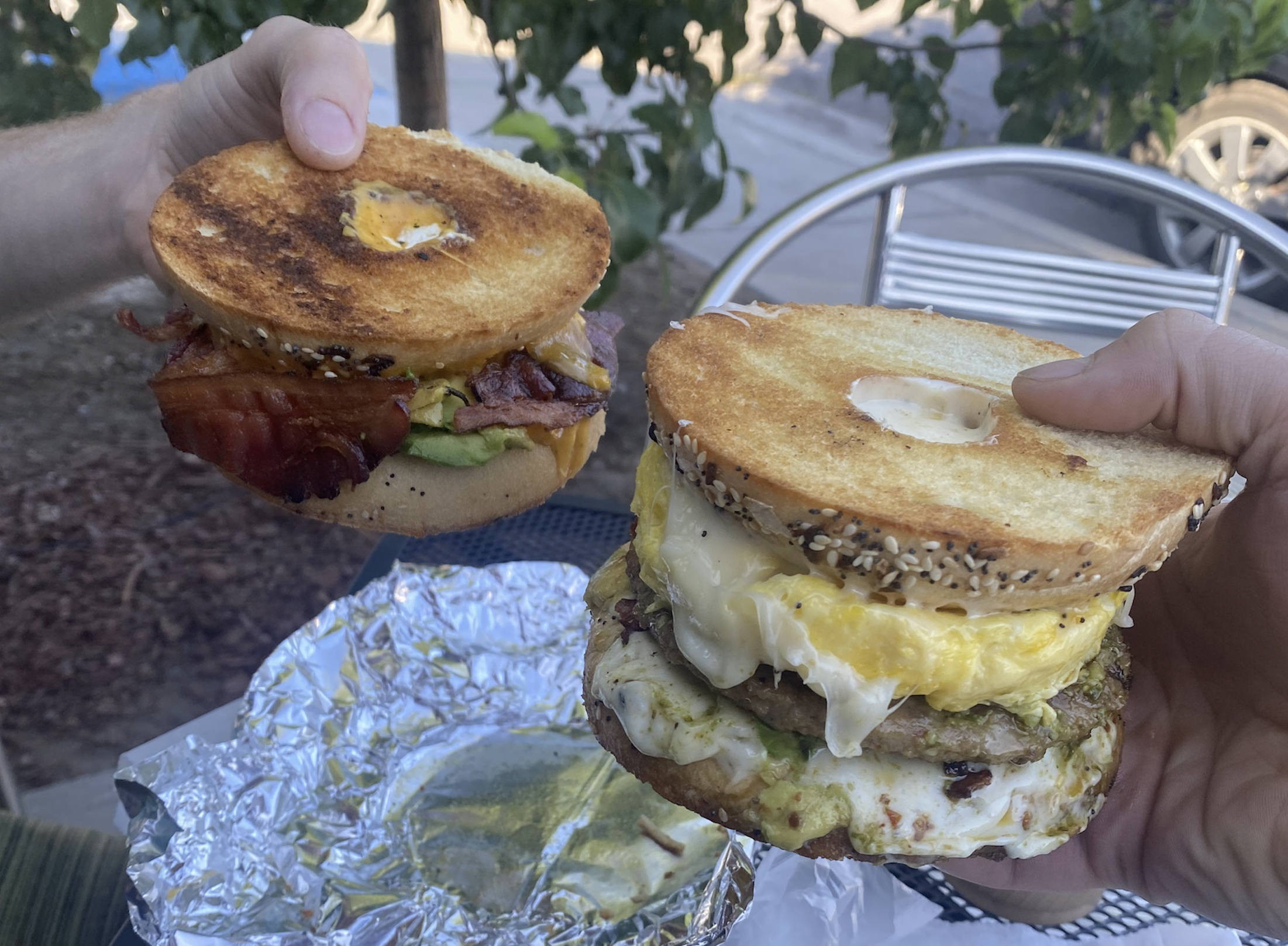 Two hands hold breakfast sandwiches made with bagels, eggs, bacon, avocado, cheese, and sausage. The sandwiches are toasted, and there is a foil wrapper on the table outdoors.