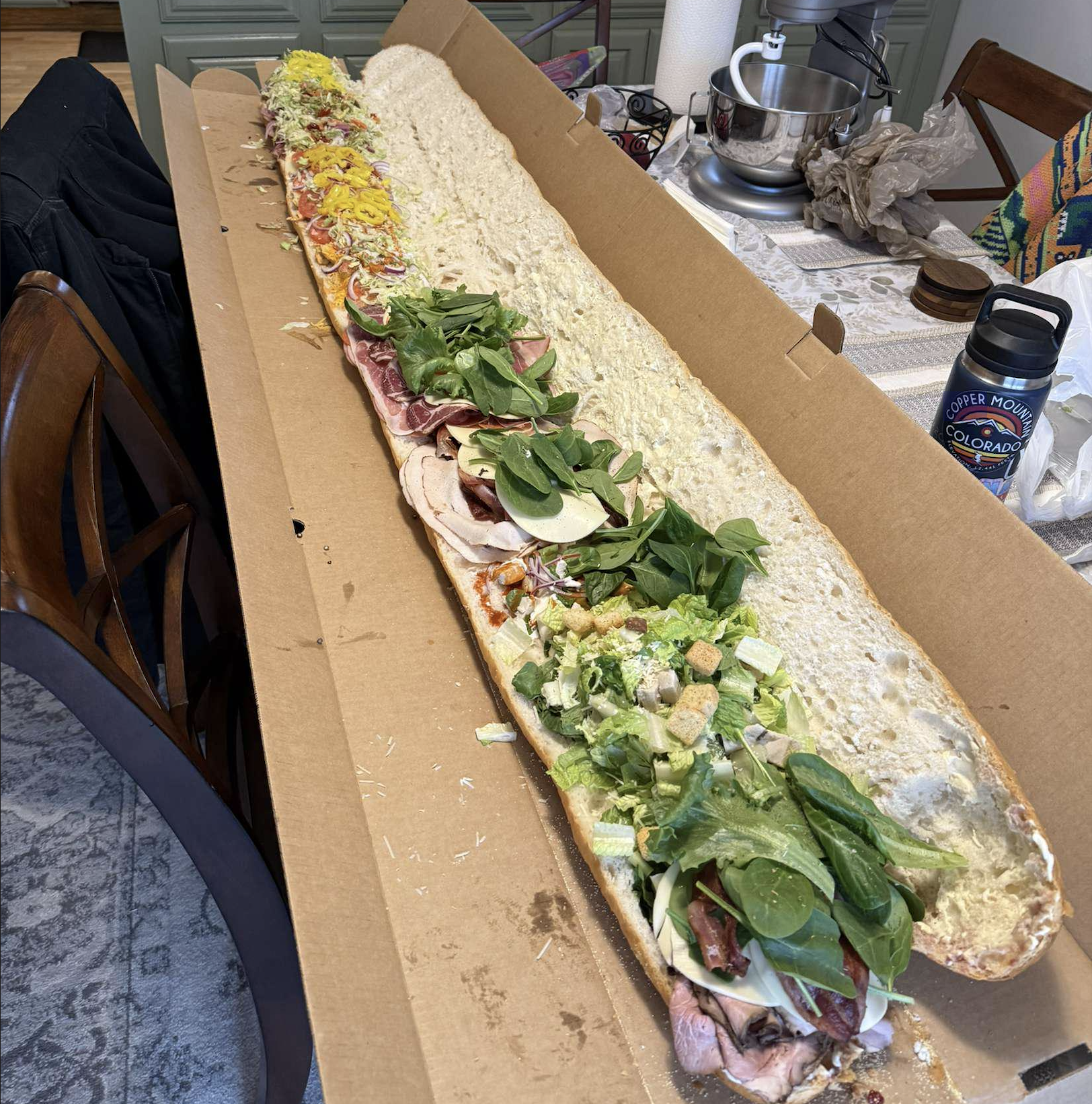 A very long sandwich in an open box sits on a table, filled with various meats, cheeses, lettuce, spinach, and other toppings, with kitchen items and a water bottle nearby.