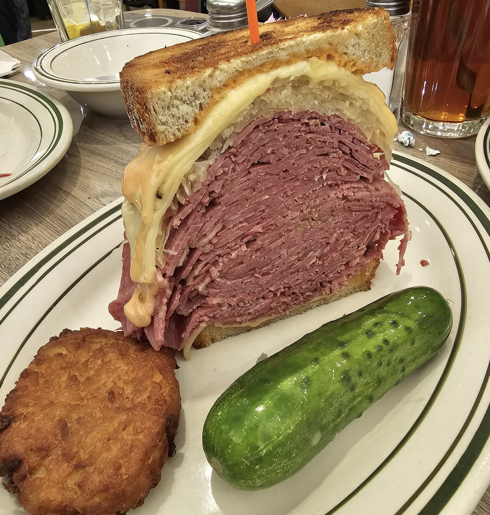 A thick pastrami sandwich with melted cheese and sauerkraut is served on a plate with a crispy potato pancake and a whole pickle.