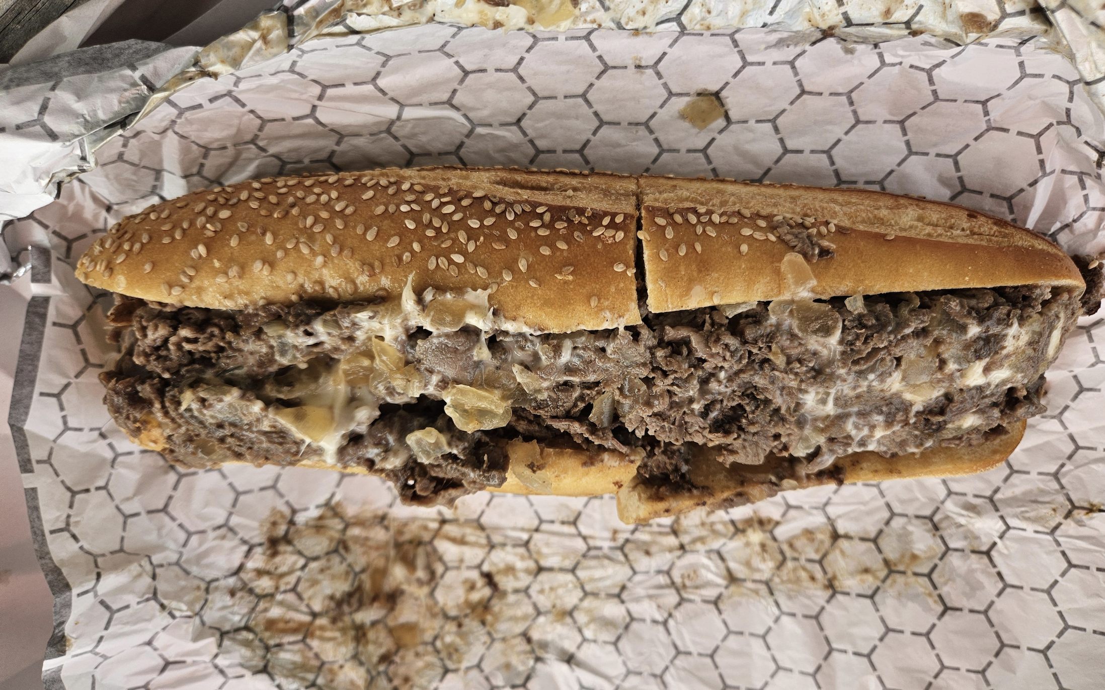A cheesesteak sandwich with melted cheese and chopped steak on a sesame seed hoagie roll, sliced in half, resting on deli paper with some spilled filling.