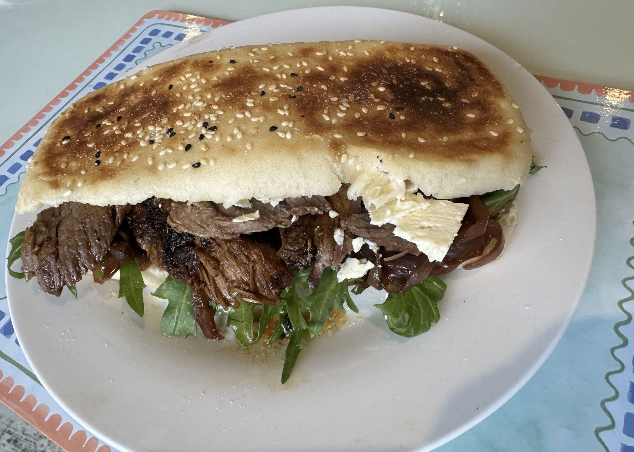 A sandwich with grilled meat, arugula, caramelized onions, and white cheese on a toasted sesame seed bun sits on a white plate with a colorful border.