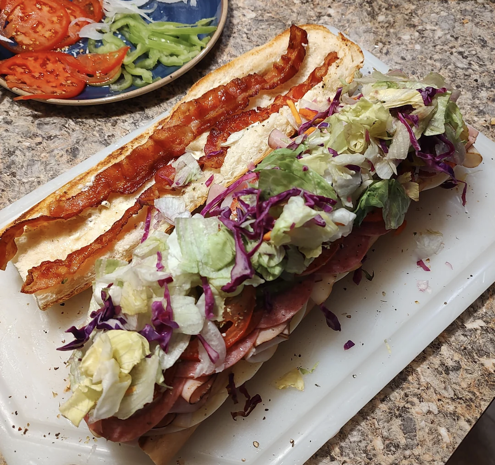 A sub sandwich on a cutting board is piled with lettuce, cabbage, meats, bacon, and cheese. Sliced tomatoes and green peppers are on a nearby plate.