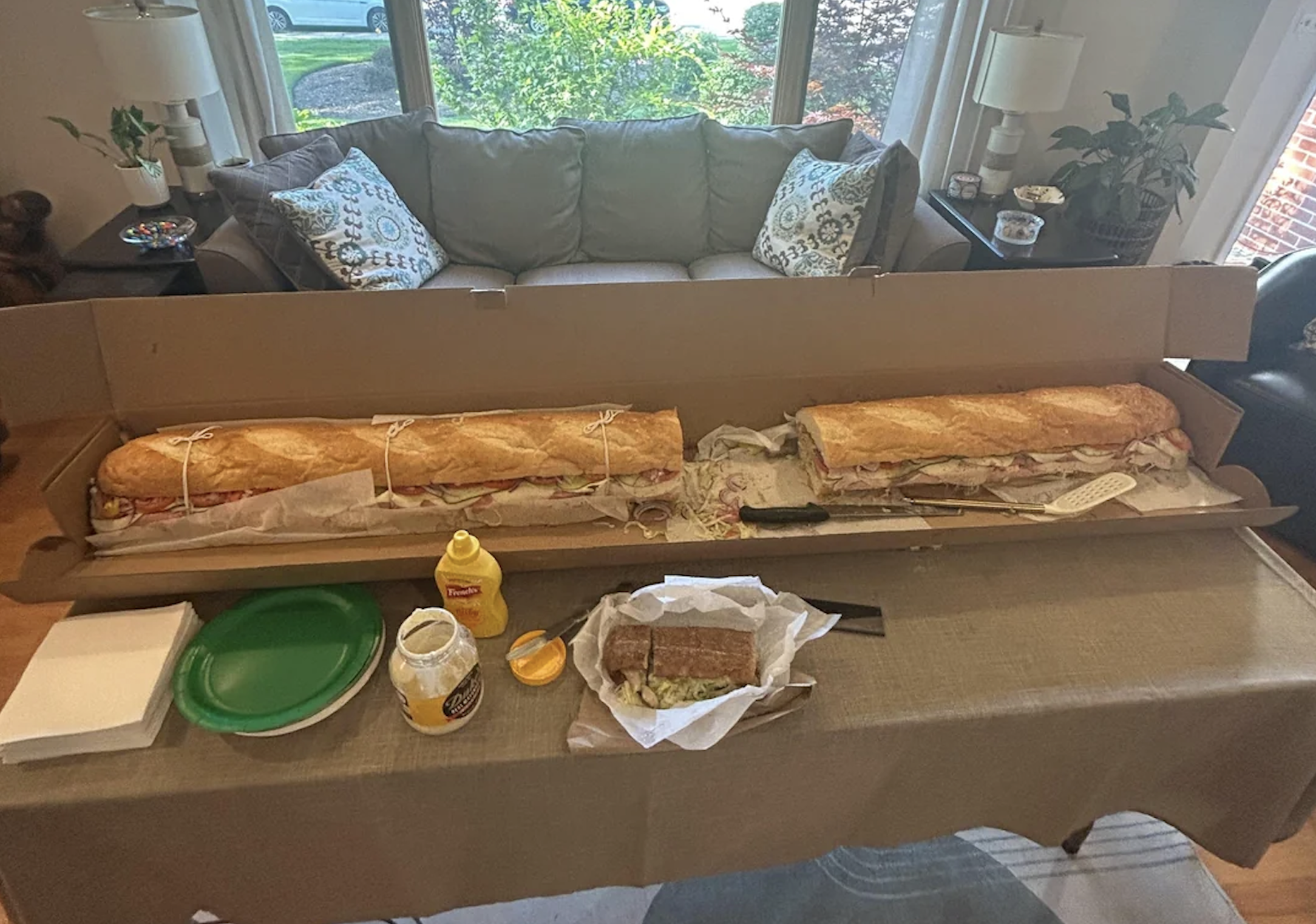 A giant sub sandwich in a long box sits on a table with plates, mustard, and a smaller wrapped sandwich in a cozy living room setting.