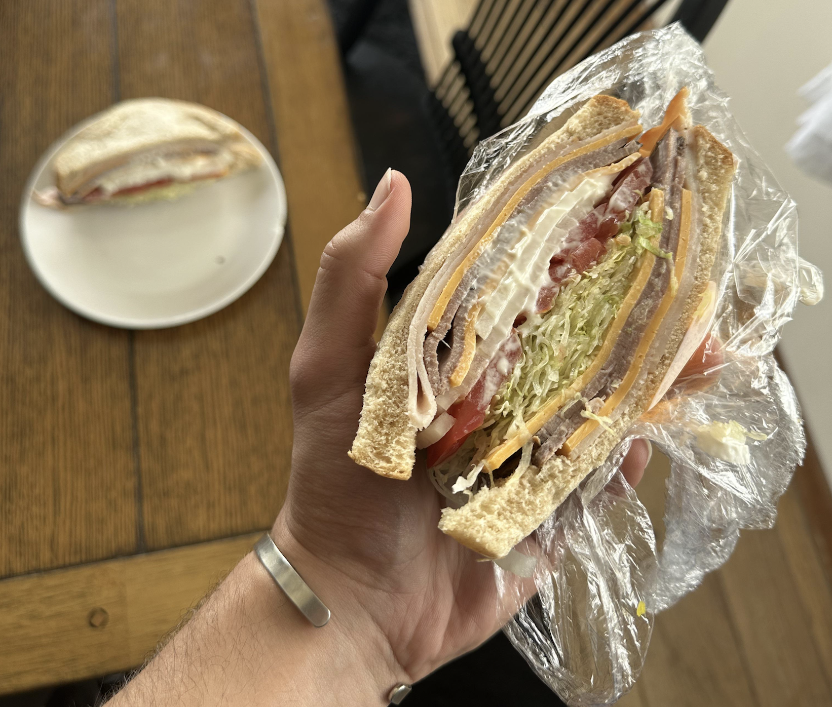 A hand holds a thick sandwich with turkey, cheddar cheese, lettuce, tomato, and mayonnaise on white bread, partially wrapped in plastic. Another sandwich half sits on a plate on a wooden table.