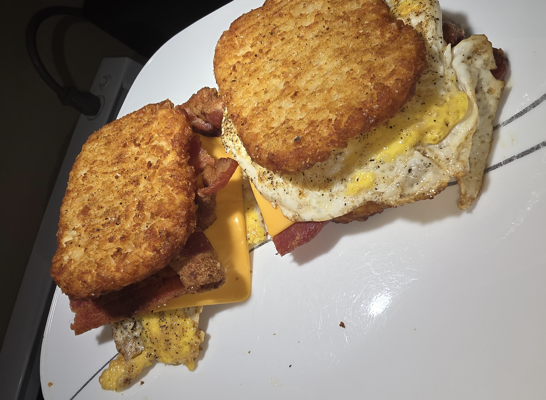 Two breakfast sandwiches with hash brown patties as buns, filled with bacon, cheddar cheese, and a fried egg, served on a white plate.