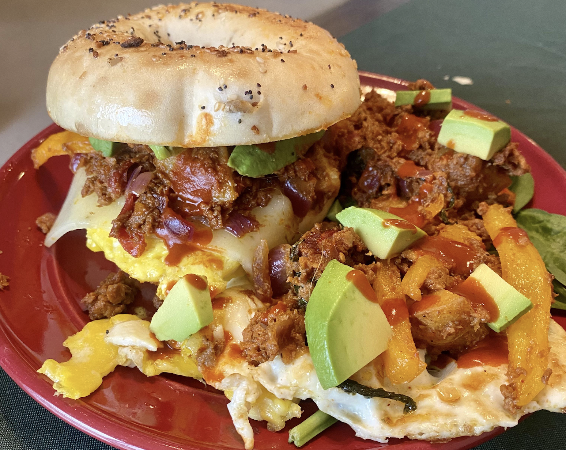 A bagel sandwich with eggs, chorizo, avocado chunks, bell peppers, spinach, and hot sauce sits on a red plate. The sandwich is open-faced with ingredients spilling out.