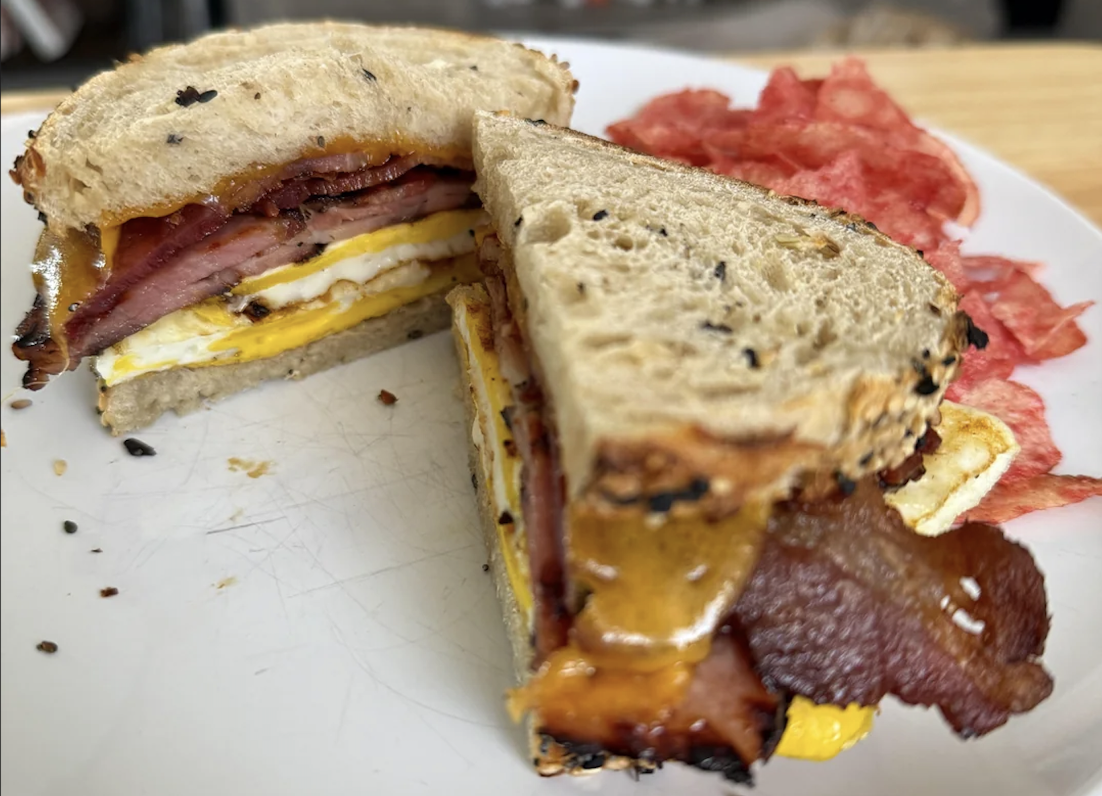 A sandwich with egg, bacon, and melted cheese on rye bread sits on a white plate, with a side of potato chips in the background.