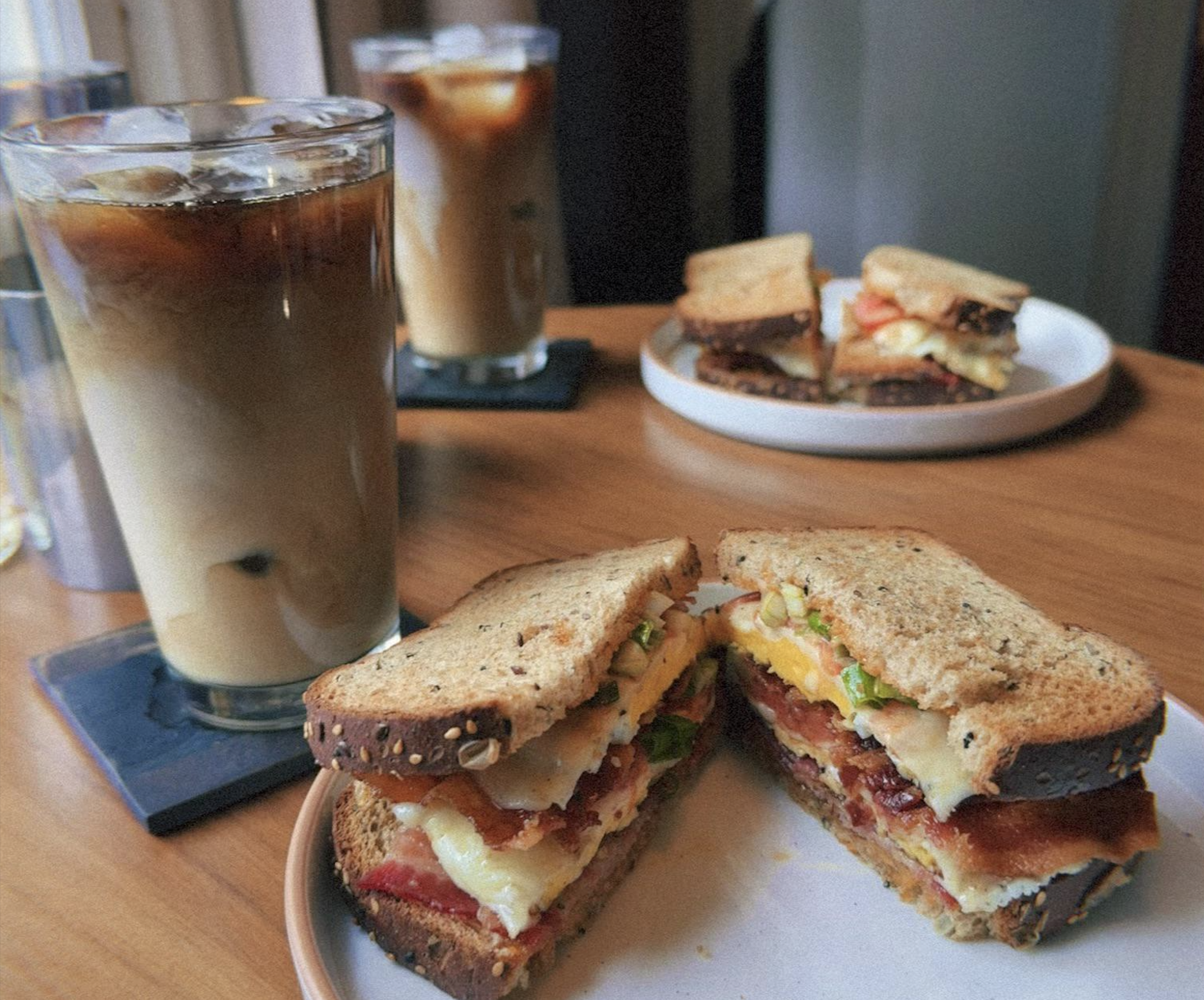 Two plates of sandwiches with bacon, egg, cheese, and greens on multigrain bread sit on a wooden table, alongside two glasses of iced coffee with milk.