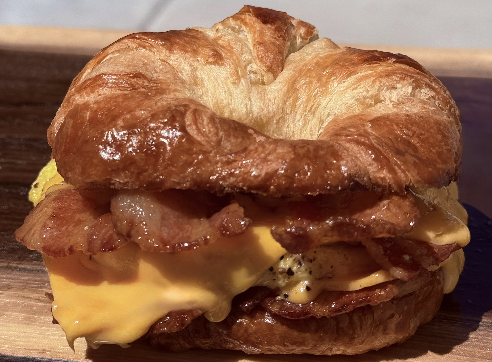 A croissant sandwich with crispy bacon, melted cheddar cheese, and scrambled eggs, served on a wooden board.