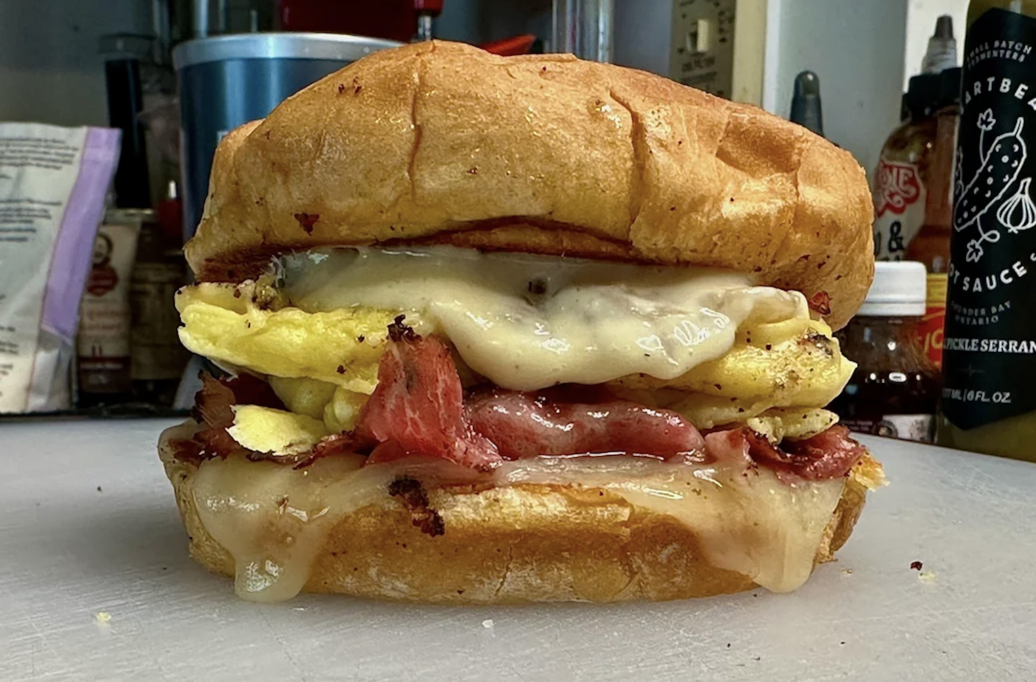A sandwich with melted cheese, scrambled eggs, and slices of meat on a toasted bun, with sauce dripping out, sits on a countertop with bottles and kitchen items in the background.