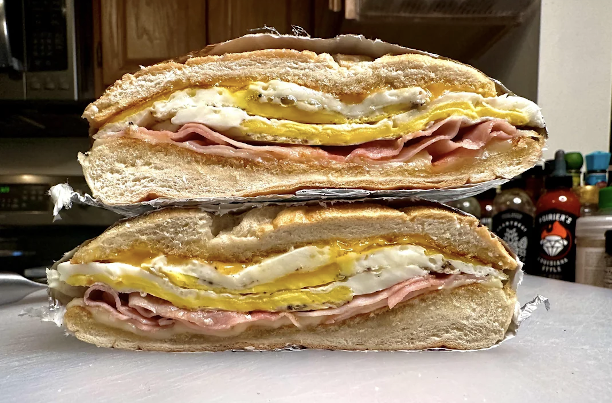 A close-up of a breakfast sandwich cut in half, showing layers of egg, ham, and melted cheese inside a thick, sliced bread roll, with bottles and a kitchen background.