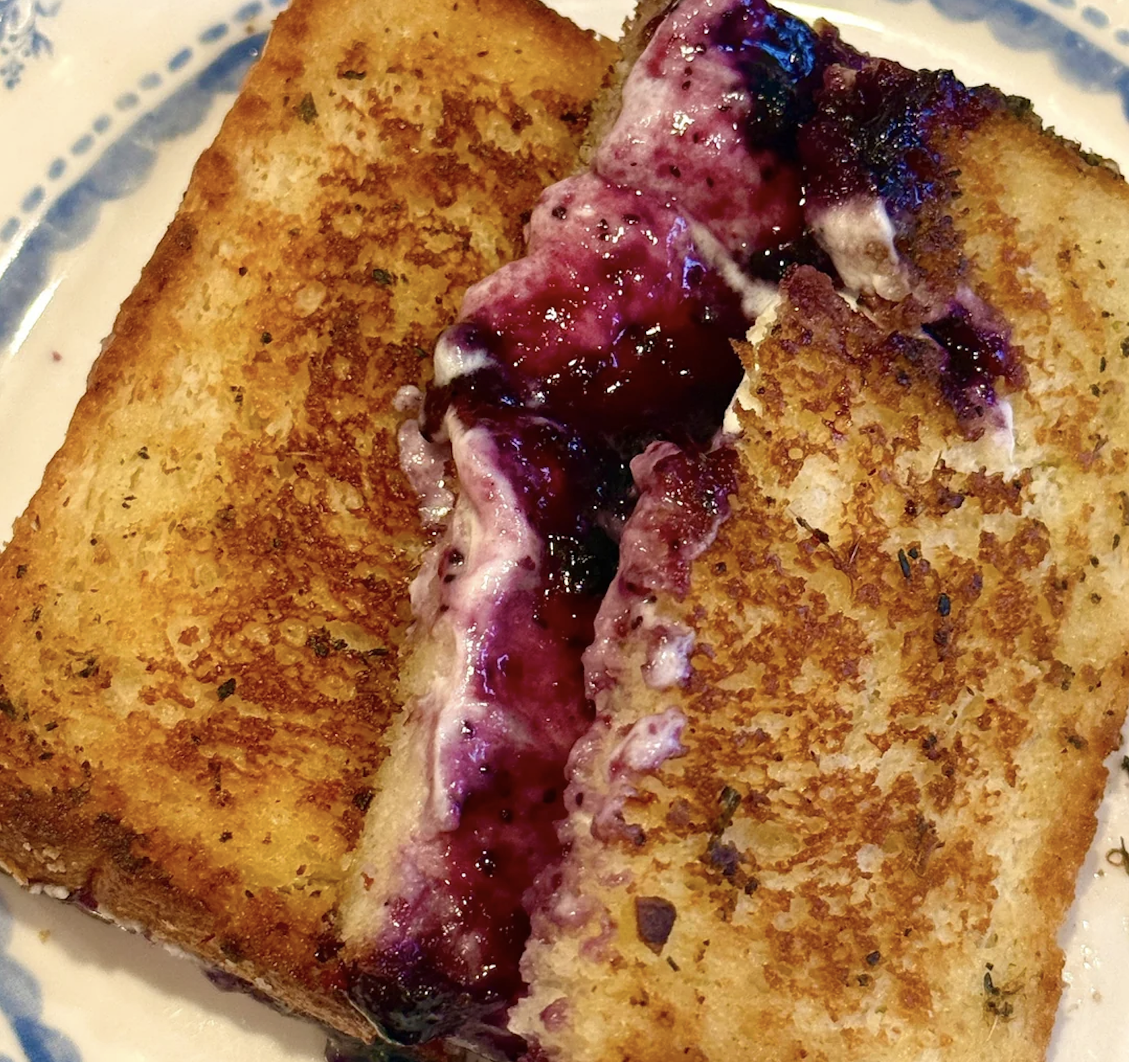 A grilled sandwich cut in half, filled with creamy cheese and dark berry jam, shown on a white plate with a blue floral pattern.
