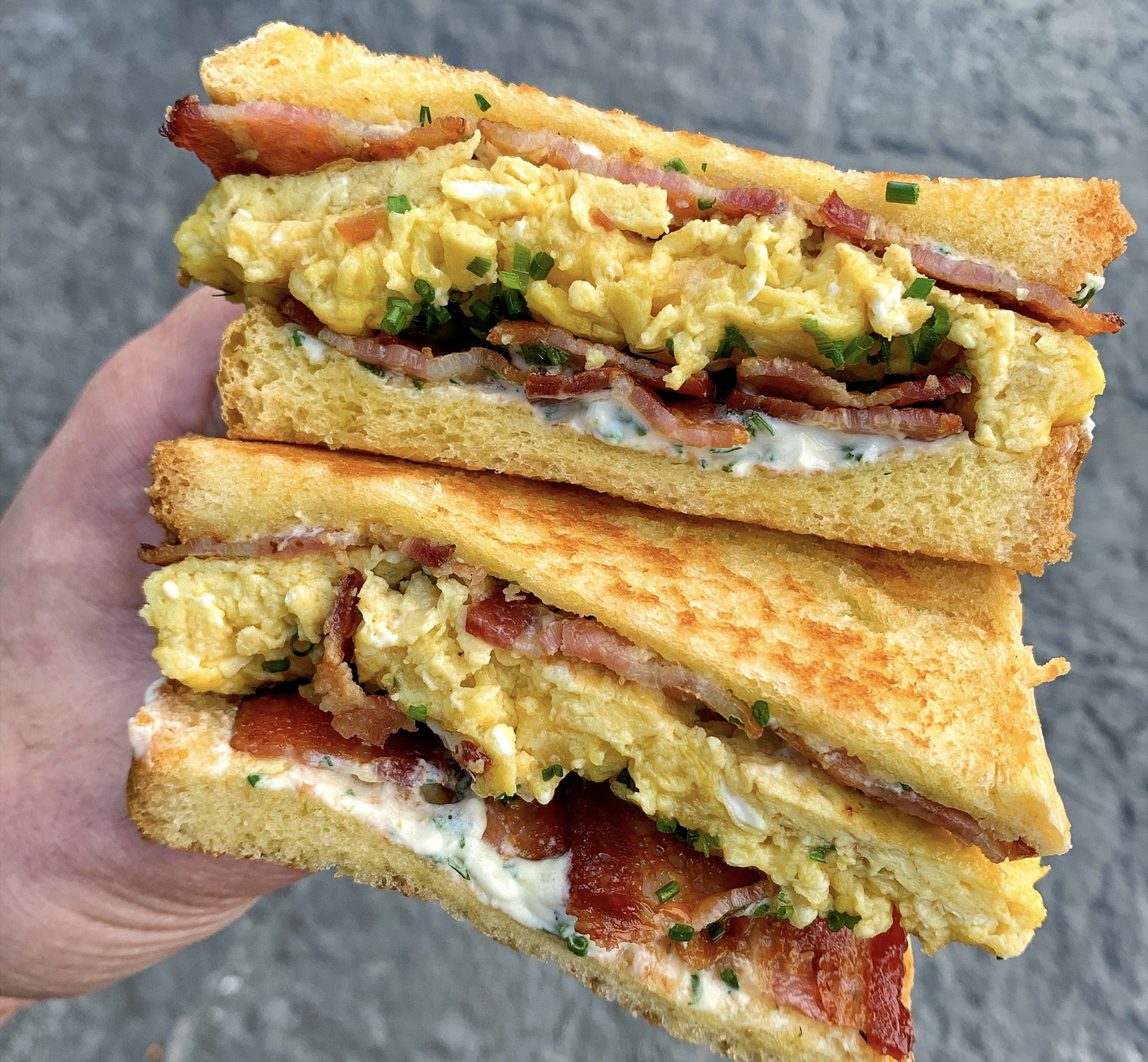 A hand holds two stacked halves of a breakfast sandwich filled with scrambled eggs, crispy bacon, chopped chives, and a creamy spread on toasted golden bread.