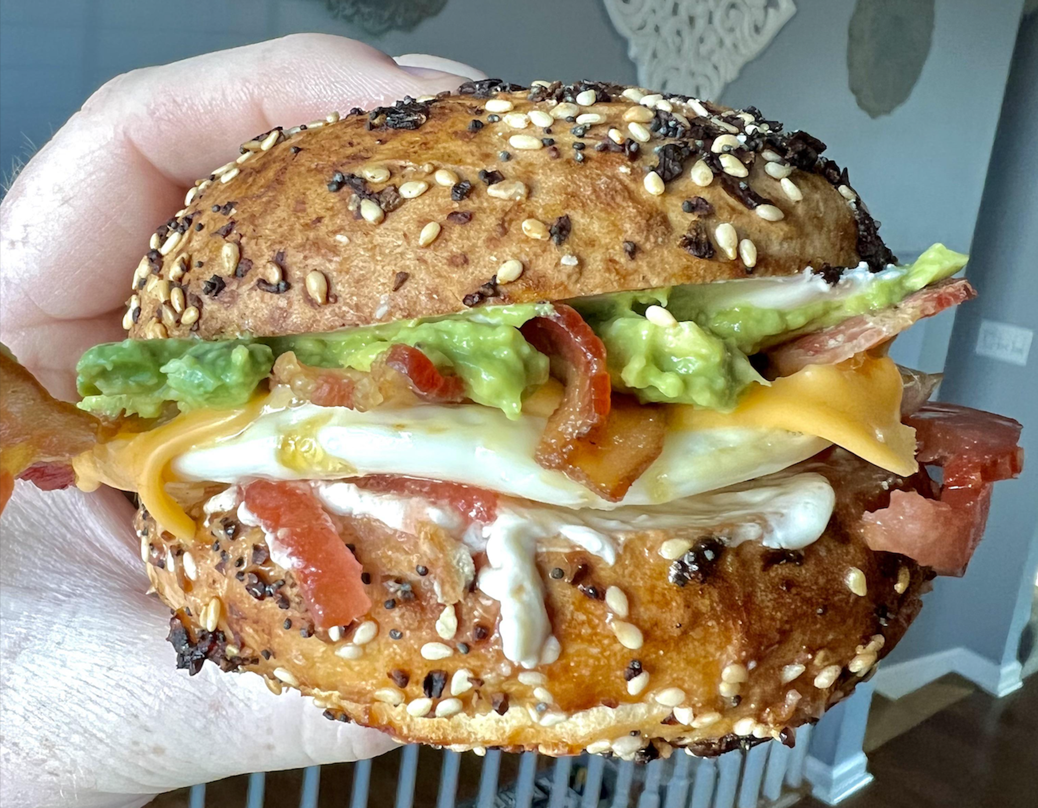 A hand holds a sandwich with a seeded bun, bacon, cheddar cheese, egg, guacamole, tomato, and mayonnaise. The background shows a light-colored room with decor on the wall.